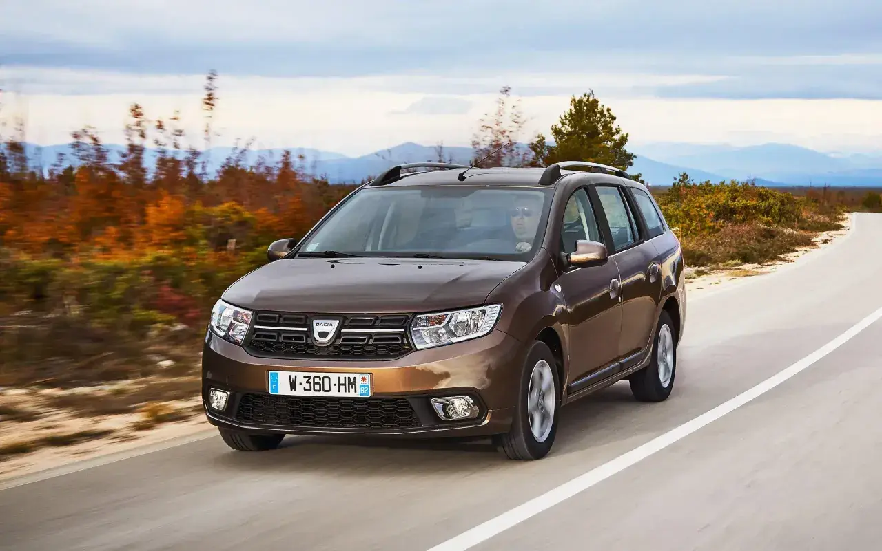 Brązowy Dacia Logan MCV jedzie po krętej drodze wśród jesiennych drzew i gór w tle.