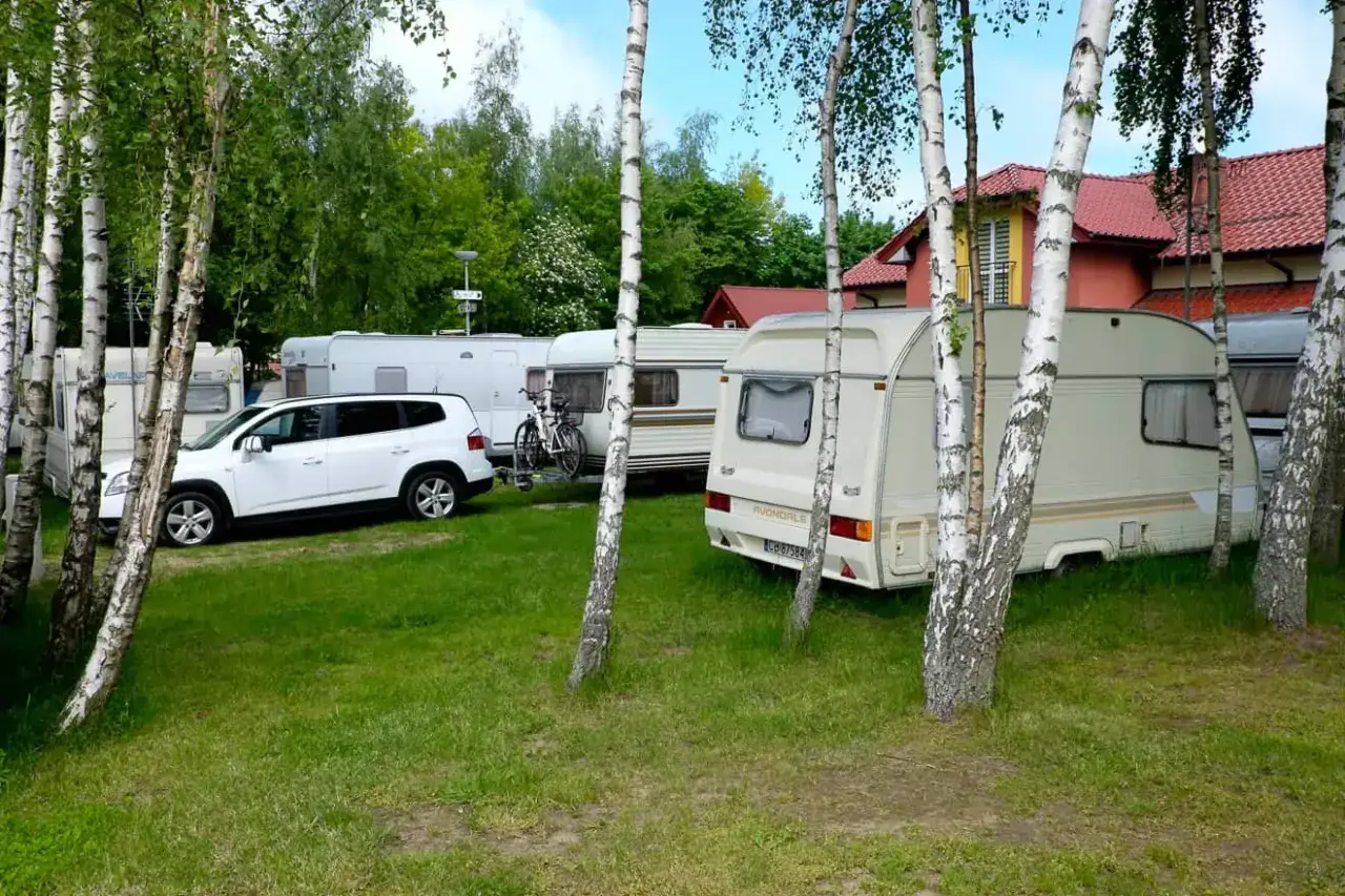 Wesoły camp Polska z przyczepami kempingowymi i białym autem, otoczony drzewami.
