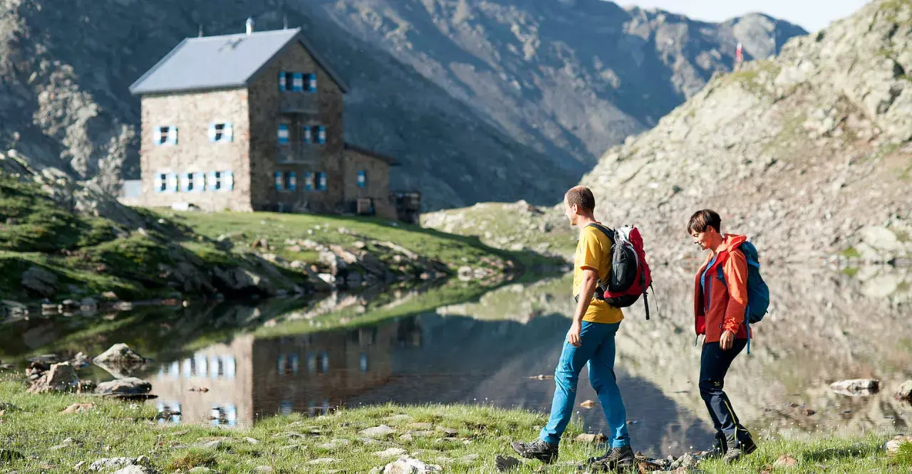 Zwei Wanderer auf einer Hufeisentour im Sarntal, vorbei an einer Berghütte und einem Bergsee.