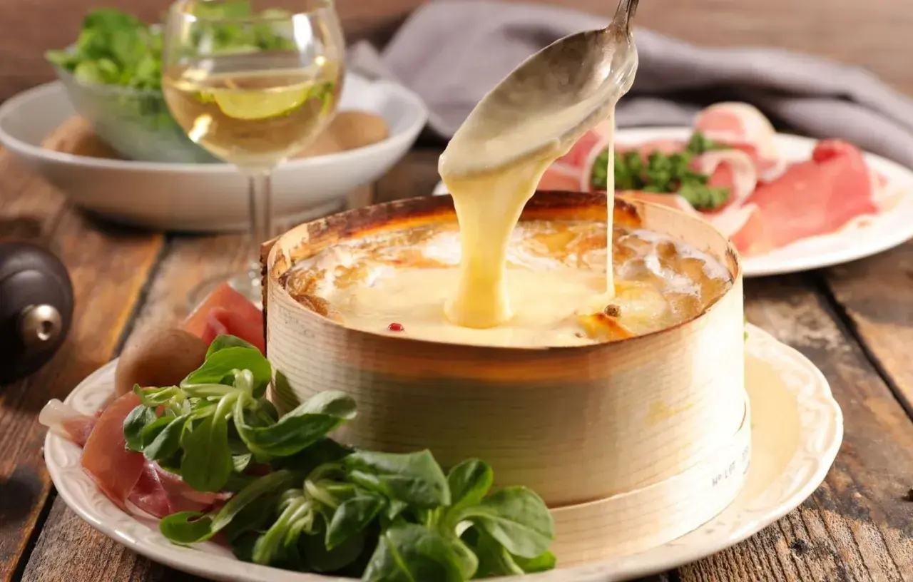 Un Mont d'Or fondant est servi avec une cuillère, accompagné de charcuterie, de pommes de terre et d'une salade verte. Un verre de vin blanc complète ce plat gourmand.