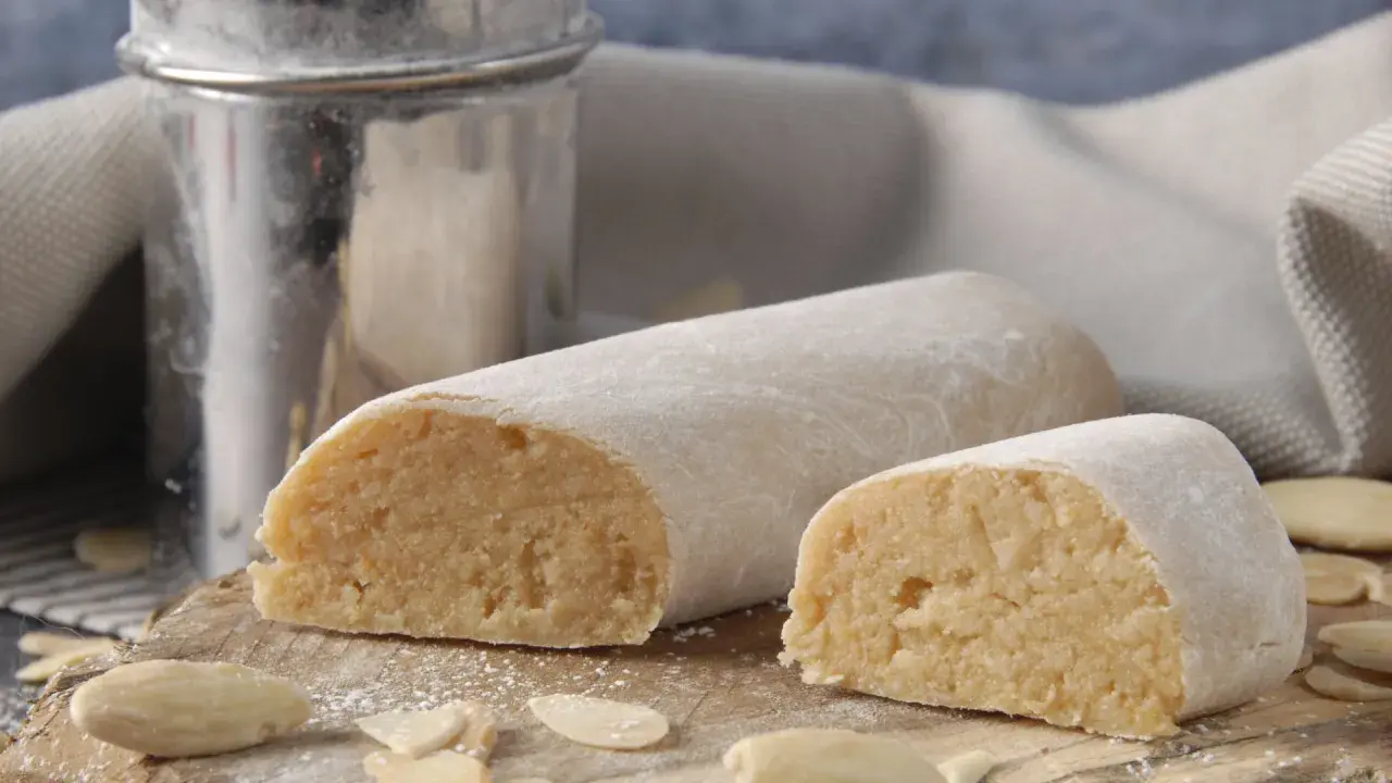 Pâte d'amande, c'est quoi ? Deux rouleaux de massepain, saupoudrés de sucre glace, sur une planche en bois avec des amandes effilées.