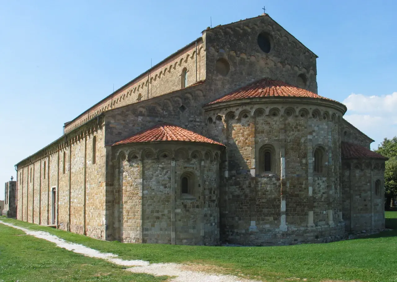 Basilica romanica di San Piero a Grado, con absidi semicircolari e tetto in tegole rosse, su un prato verde sotto un cielo azzurro.