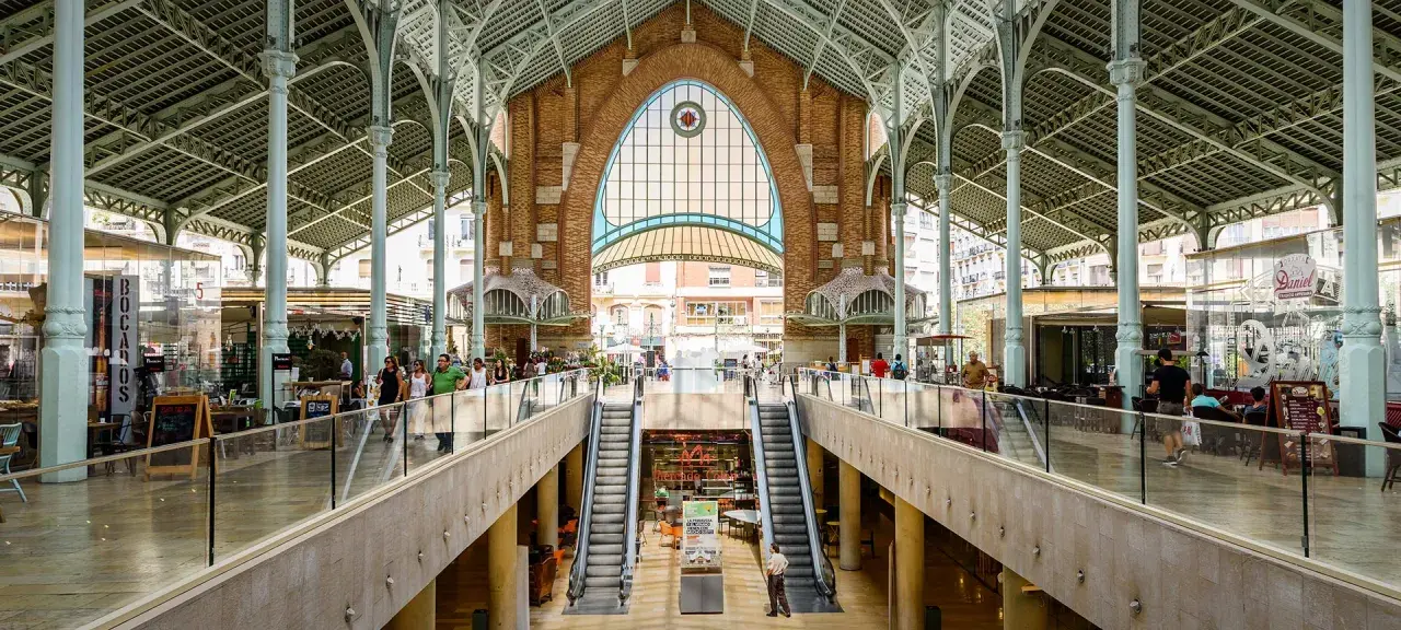 Interior del Mercado de Colón, con sus arcos de hierro y cristal, escaleras mecánicas y puestos de comida.