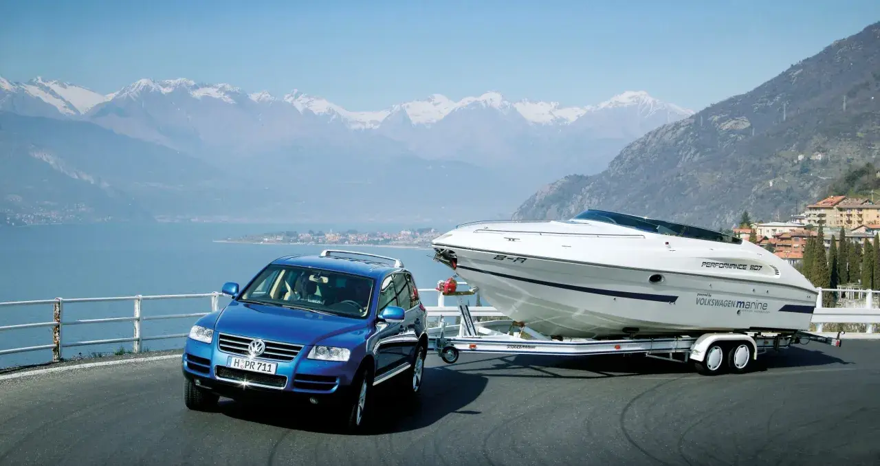 Un coche azul remolca un barco deportivo blanco por una carretera junto a un lago y montañas nevadas.