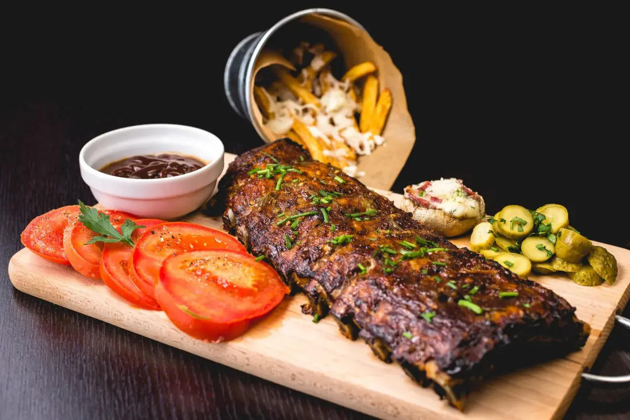 Un plat de côtes de porc au barbecue, garni de ciboulette, servi avec des tomates tranchées, des cornichons, une sauce barbecue et des frites au fromage.