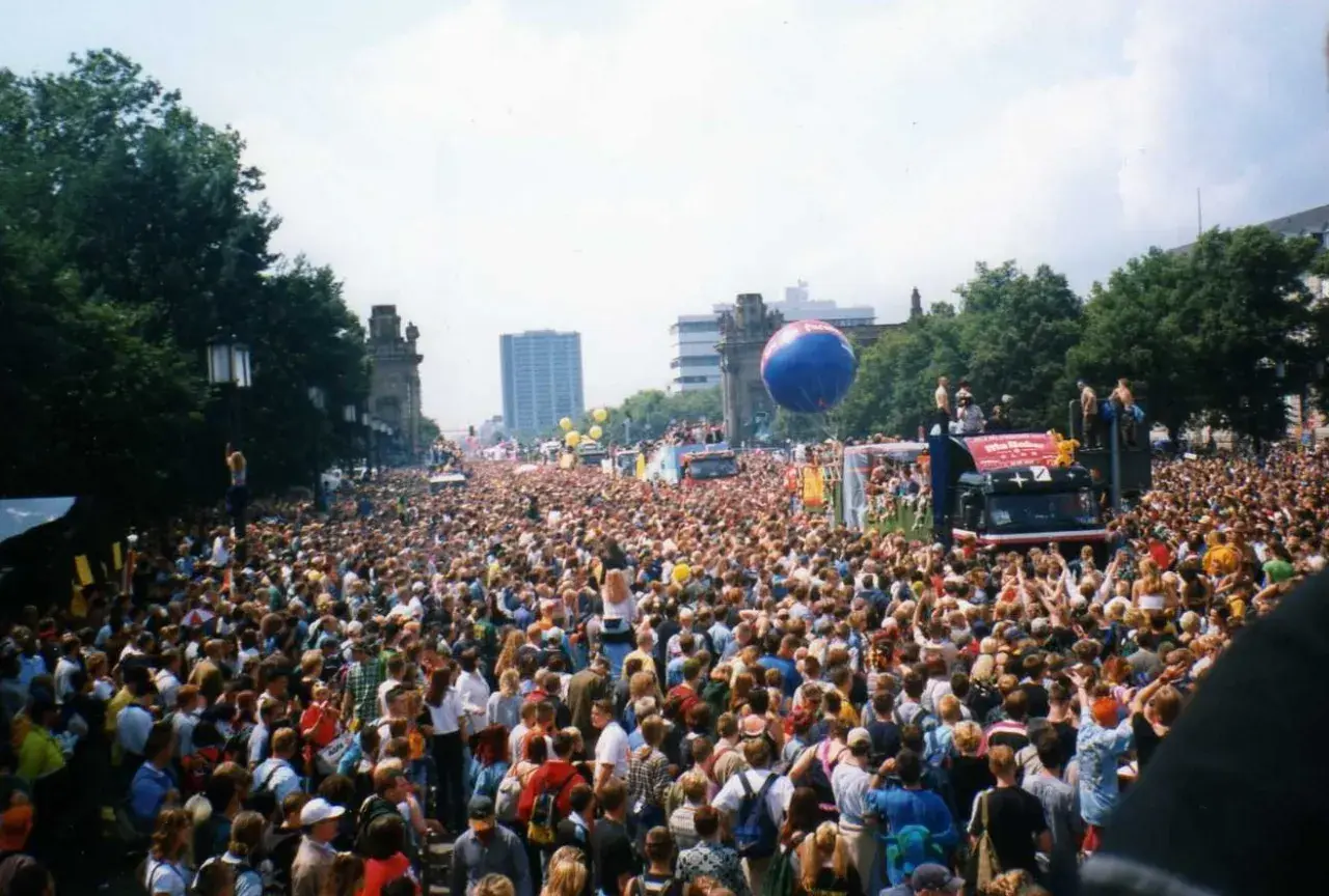 Tłum ludzi na ulicy podczas Love Parade, z wielkim niebieskim balonem i ciężarówką z platformą.