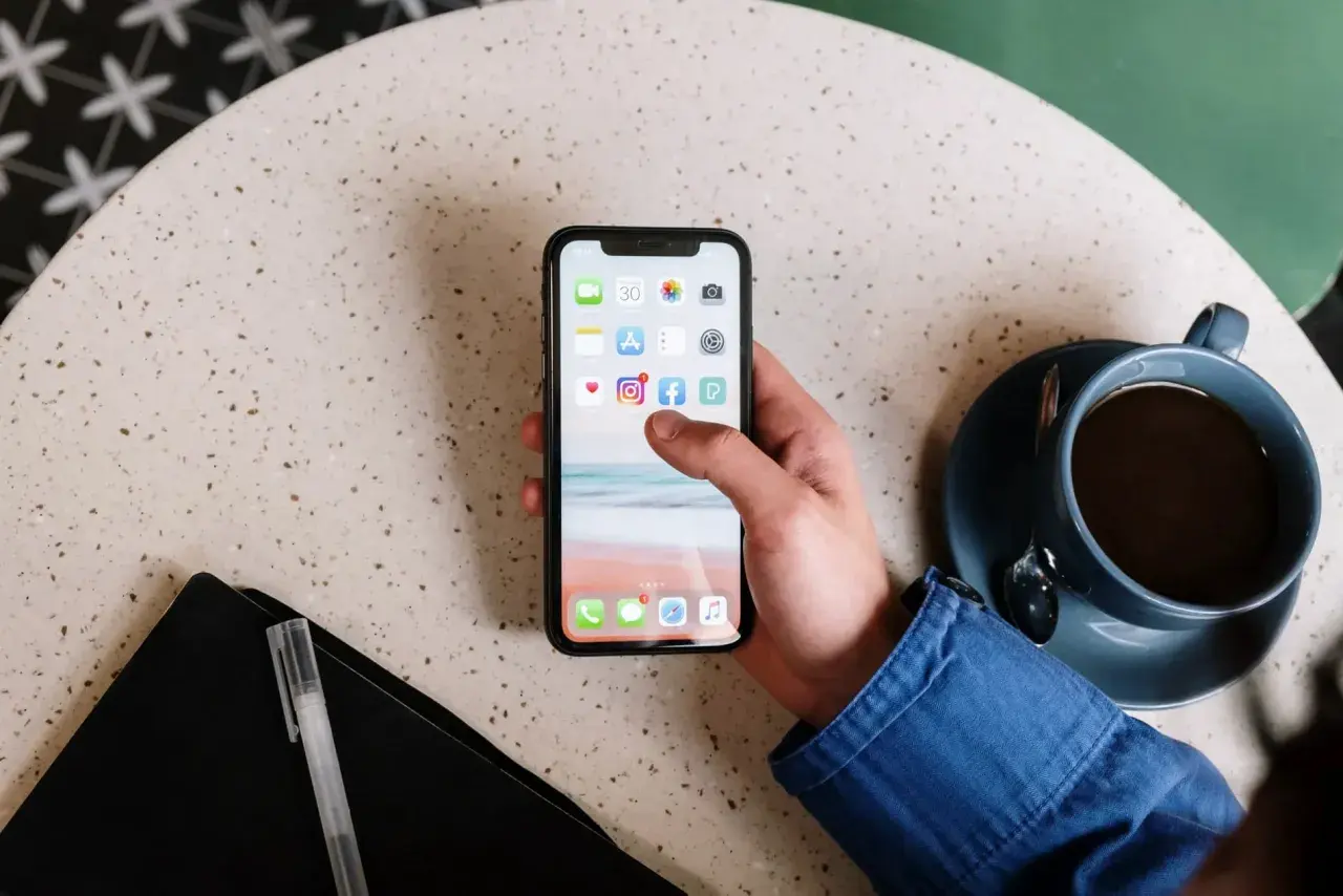 Main d'une personne tenant un smartphone affichant des icônes d'applications, comme un exemple d'application web. Une tasse de café est à côté.