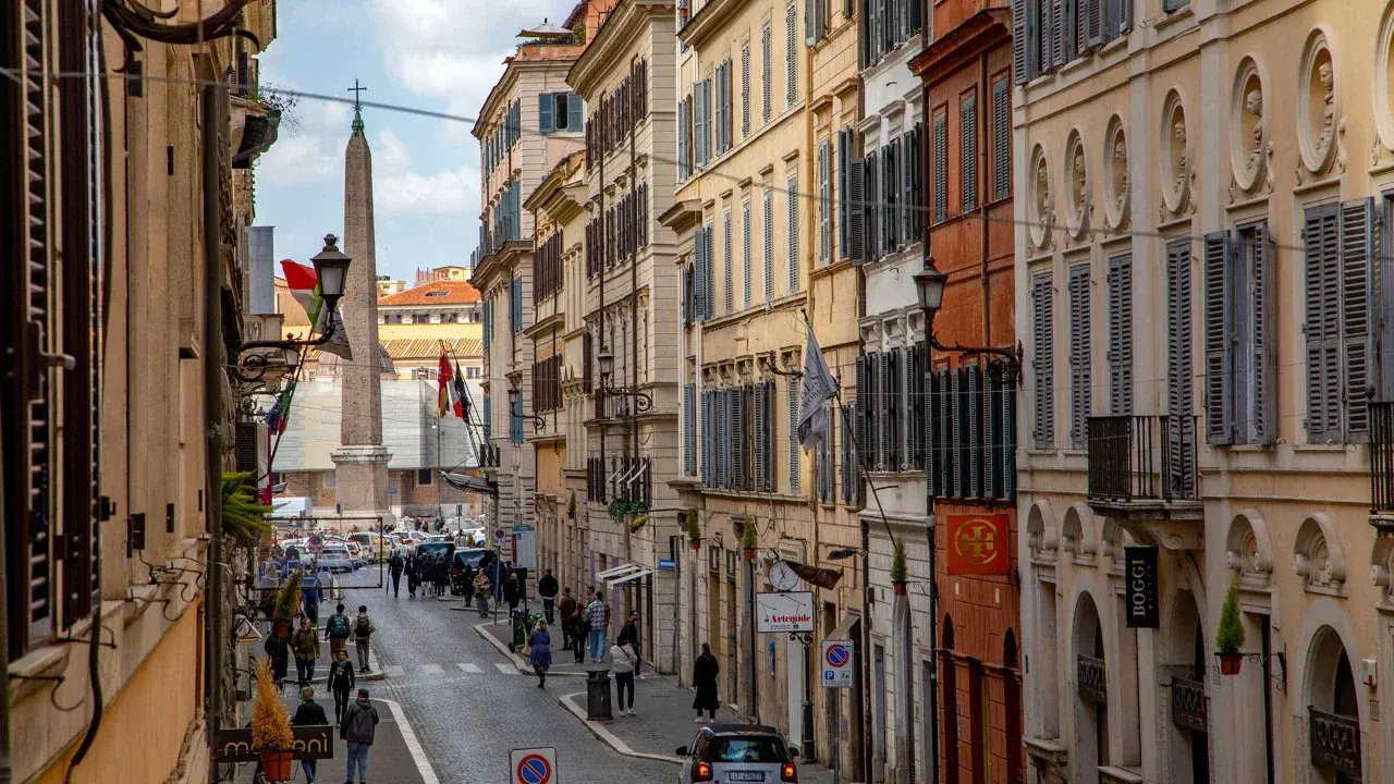 Una strada romana con palazzi storici, un obelisco e gente che passeggia. Si intuisce quanto è lunga Via del Corso, con la sua prospettiva che si perde all'orizzonte.