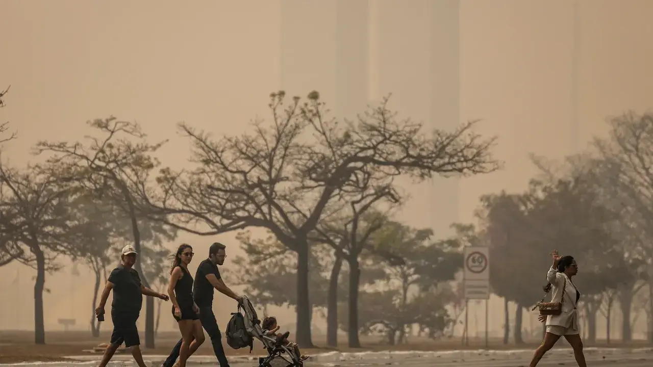 Gente camina bajo un cielo anaranjado, reflejo de las causas de la contaminación del aire. Árboles secos y edificios difuminados completan la escena.
