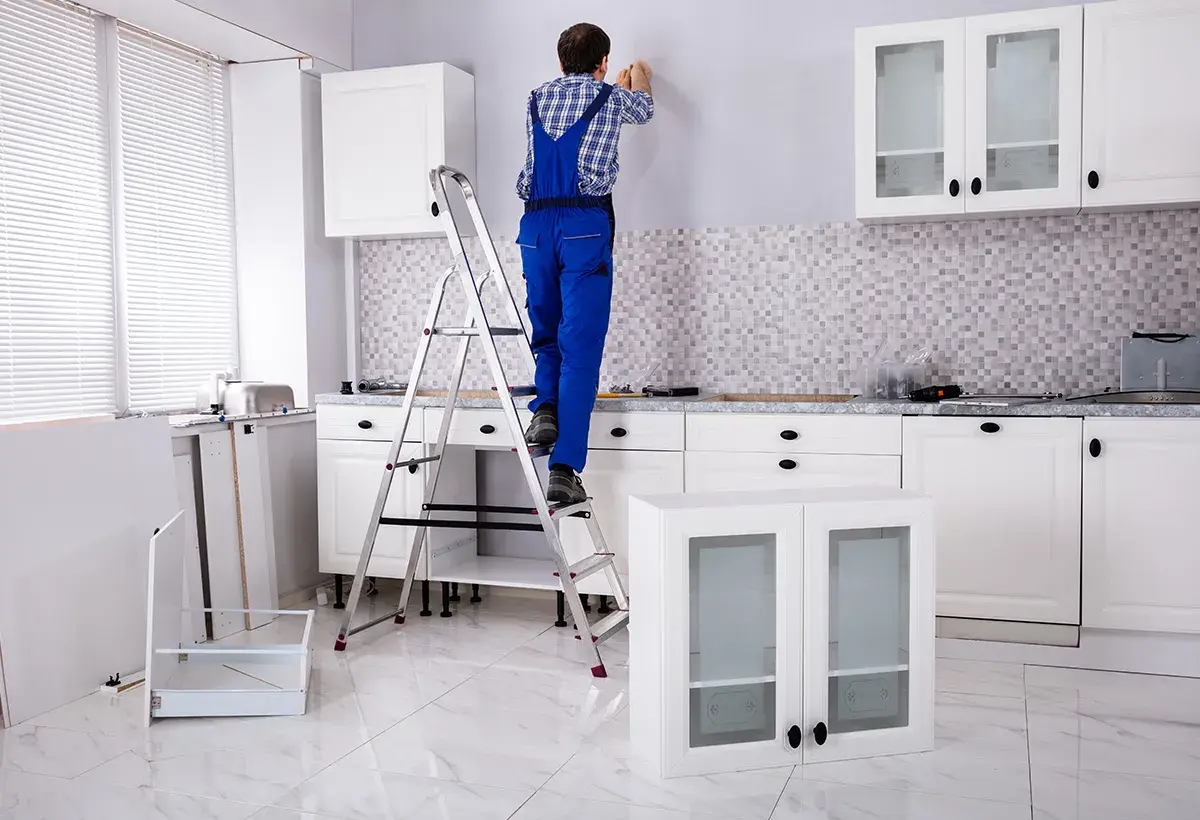 Un homme en salopette bleue est sur un escabeau, prêt à fixer un meuble au mur. La cuisine est en cours d'installation.