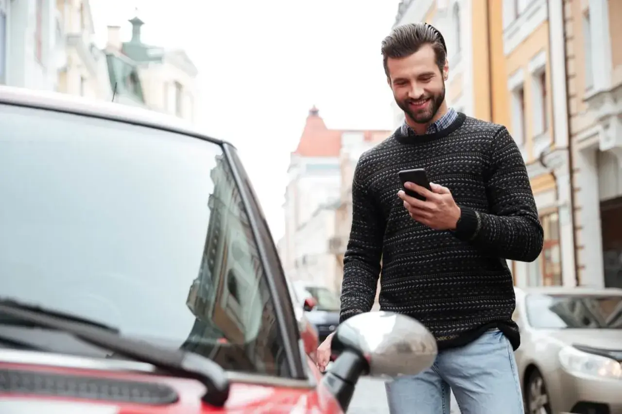 Uśmiechnięty mężczyzna z telefonem w ręku stoi obok czerwonego samochodu na ulicy.