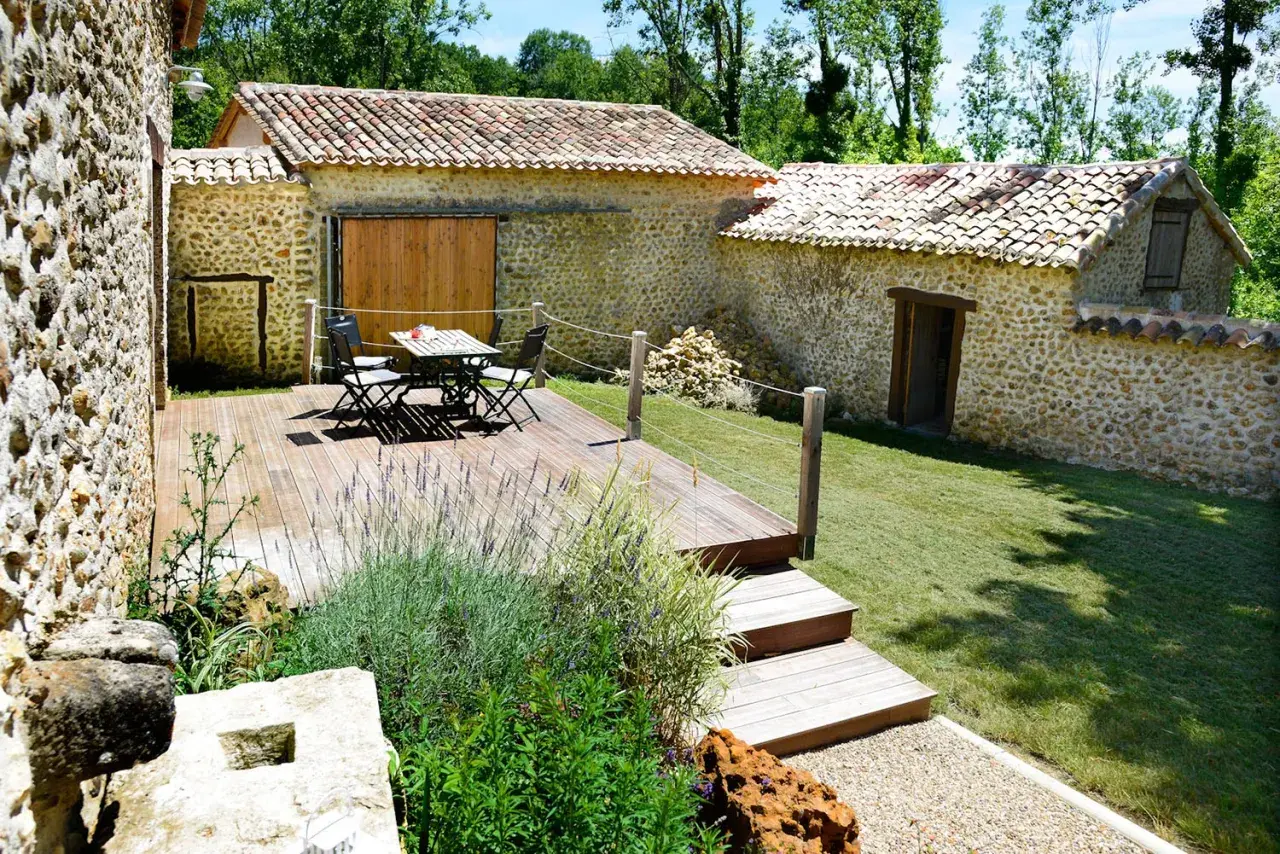 Aménagement longère : terrasse en bois avec mobilier, jardin verdoyant et murs en pierre.