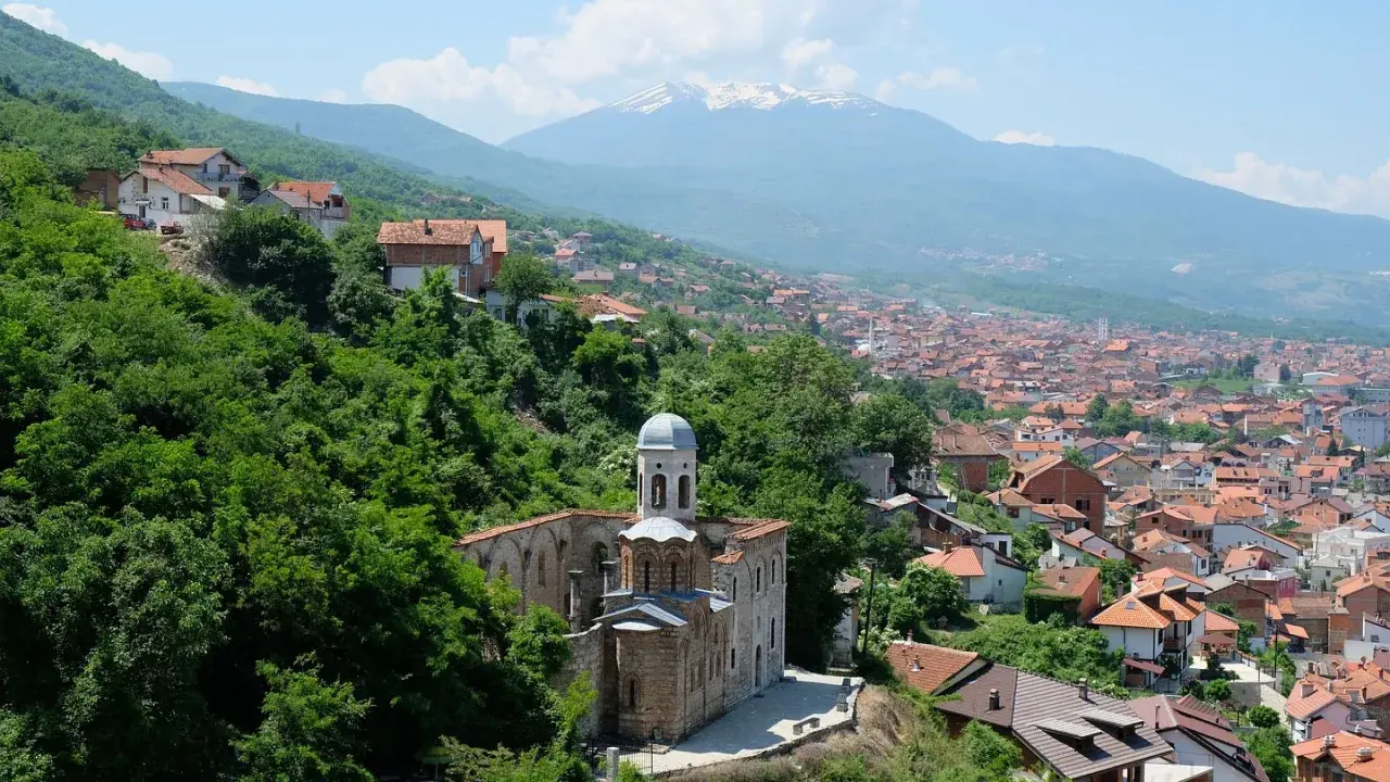 Eine malerische **Kosovo Landschaft** mit einer alten Kirche im Vordergrund, umgeben von üppigem Grün. Im Hintergrund erstreckt sich eine Stadt mit roten Dächern bis zu schneebedeckten Bergen.
