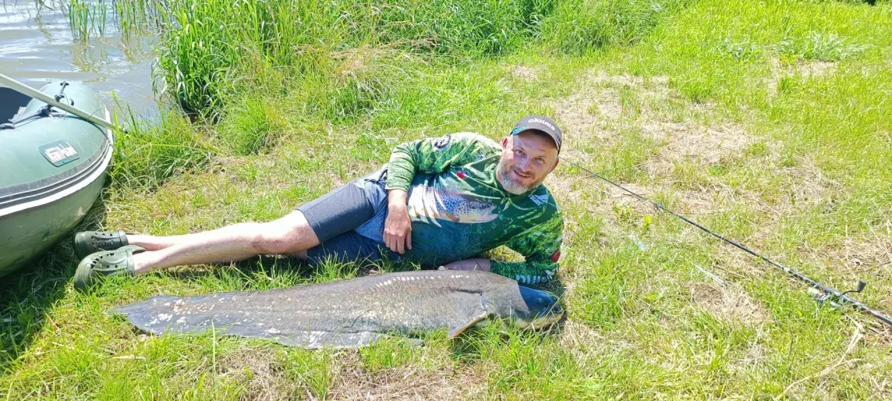 Szczęśliwy wędkarz z potężnym sumem złowionym w zbiorniku Nielisz. Obok łódka i wędka.