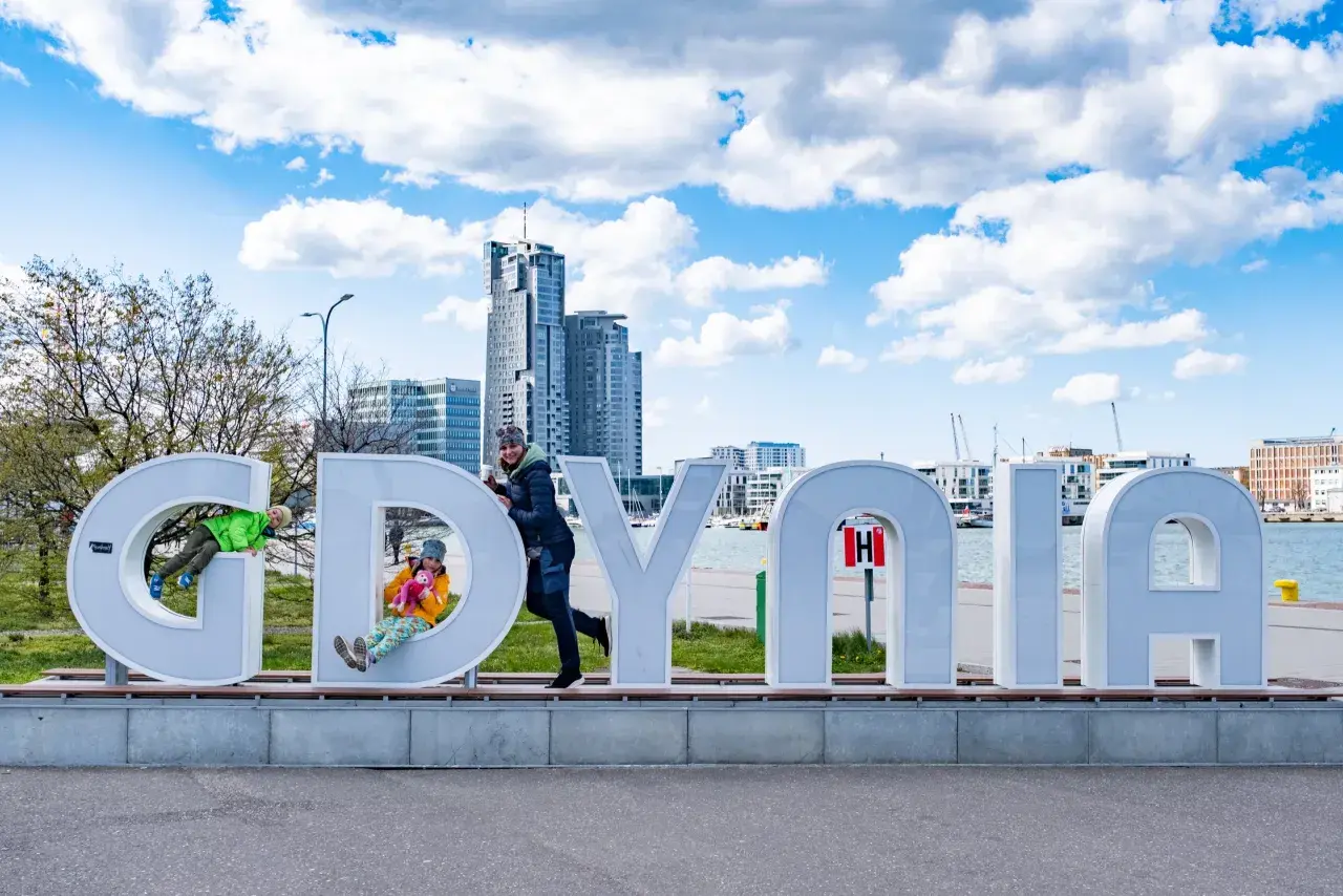 Dzieci bawią się na literach napisu "GDYNIA" na tle miejskiego krajobrazu.