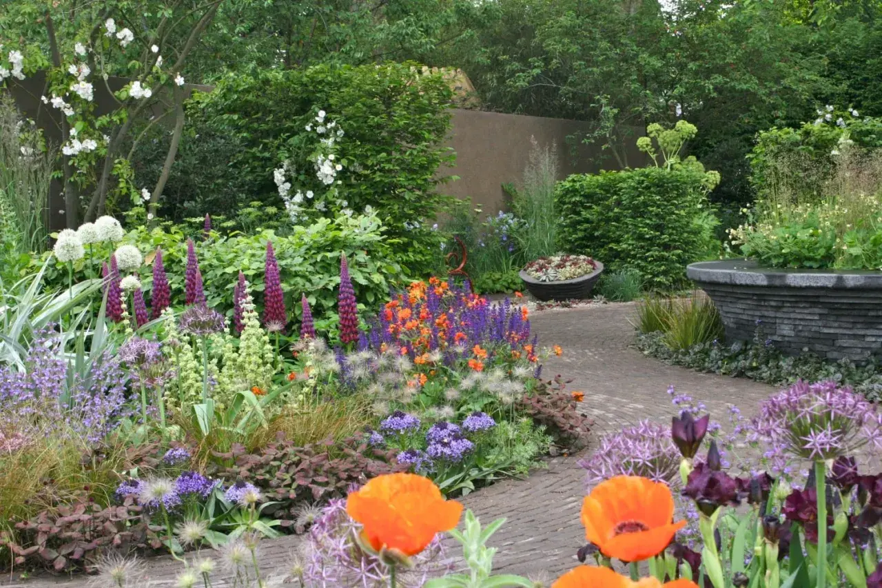 En lummig trädgård med en gång av tegelstenar. Här har någon valt att plantera blommor i en färgsprakande blandning av lila, orange och vitt.