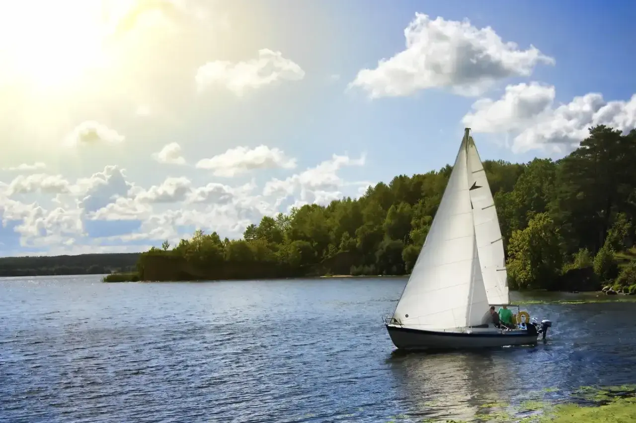 Ahoj Mazury! Biały żagiel łodzi odbija się w spokojnej tafli jeziora, otoczonej zielonymi lasami. Słońce przebija się przez chmury.