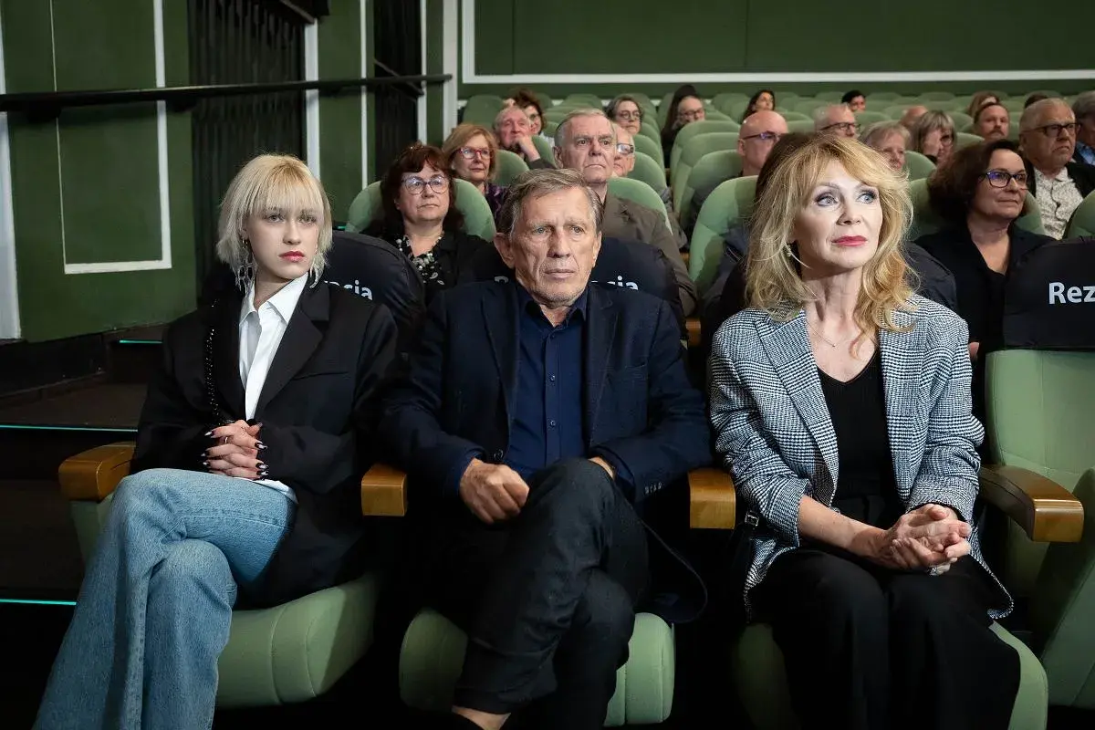 Małgorzata Englert, w wieku, siedzi w pierwszym rzędzie obok innych osób, wpatrując się w dal.