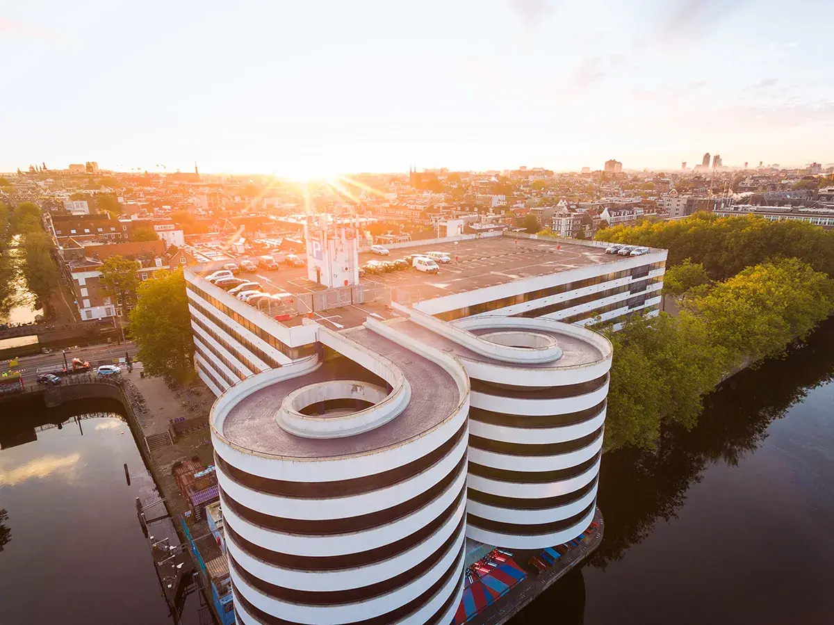 Sonnenuntergang über Amsterdam. Ein Parkhaus mit spiralförmigen Rampen ragt über den Kanal.