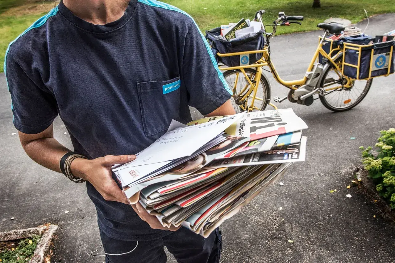 Postnord-utdelare med post i händerna, redo för postutdelning varannan dag. En gul postcykel syns i bakgrunden.