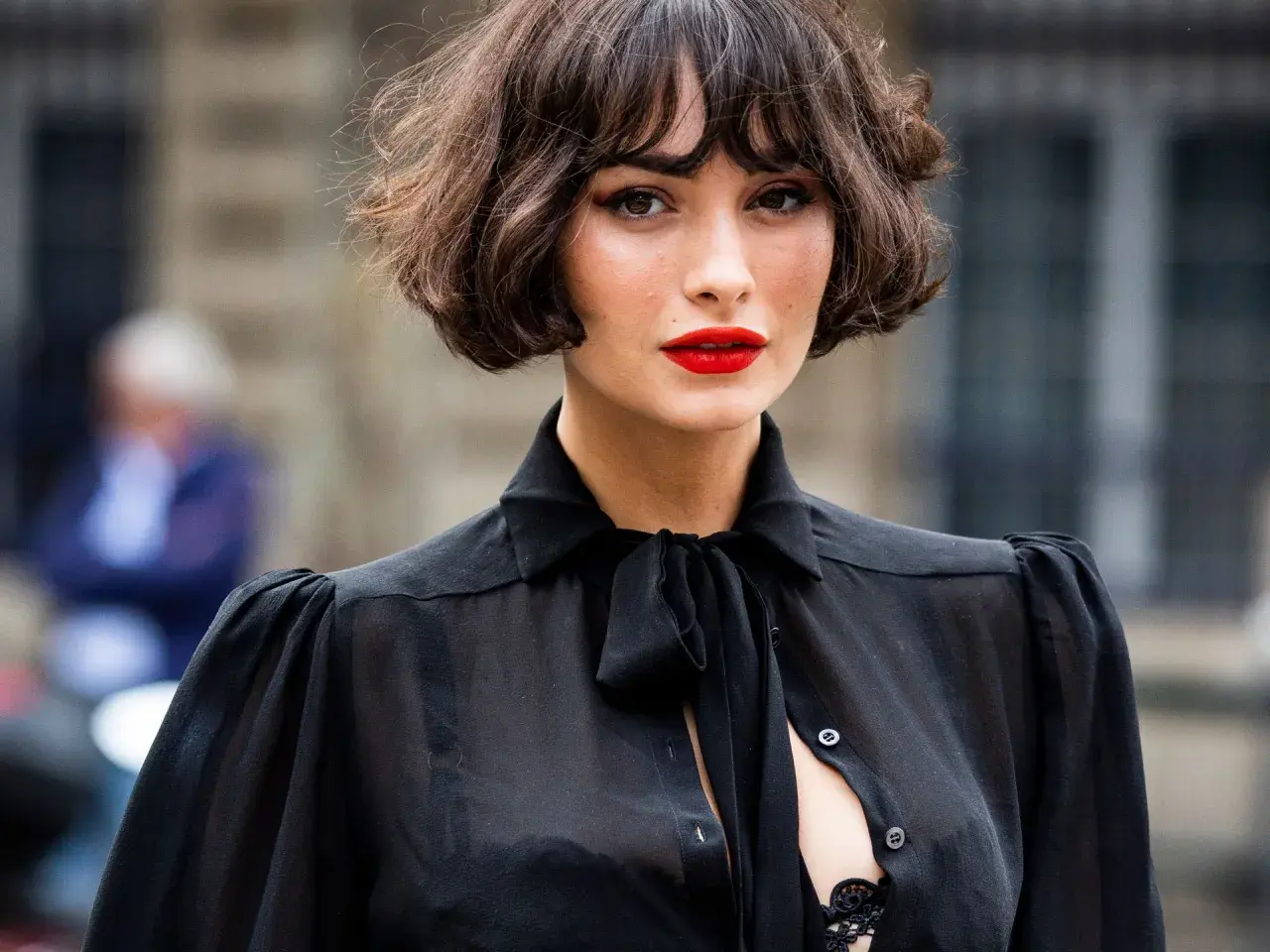 Mujer con un elegante corte de pelo **french bob**, labios rojos vibrantes y una blusa negra transparente con lazada.