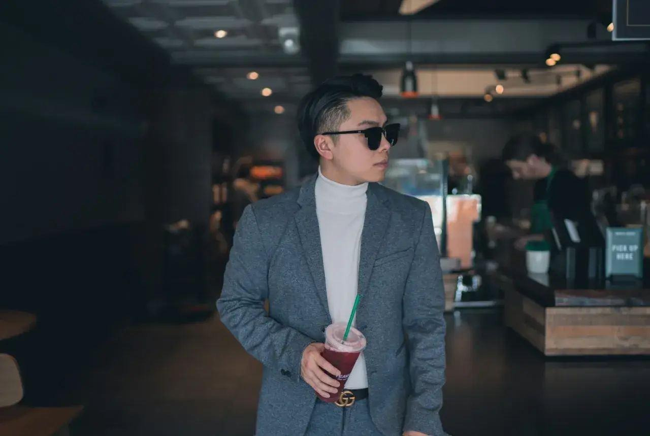 Mann im grauen Anzug mit weißem Rollkragenpullover und Sonnenbrille, hält Getränk. Das ist smart casual.