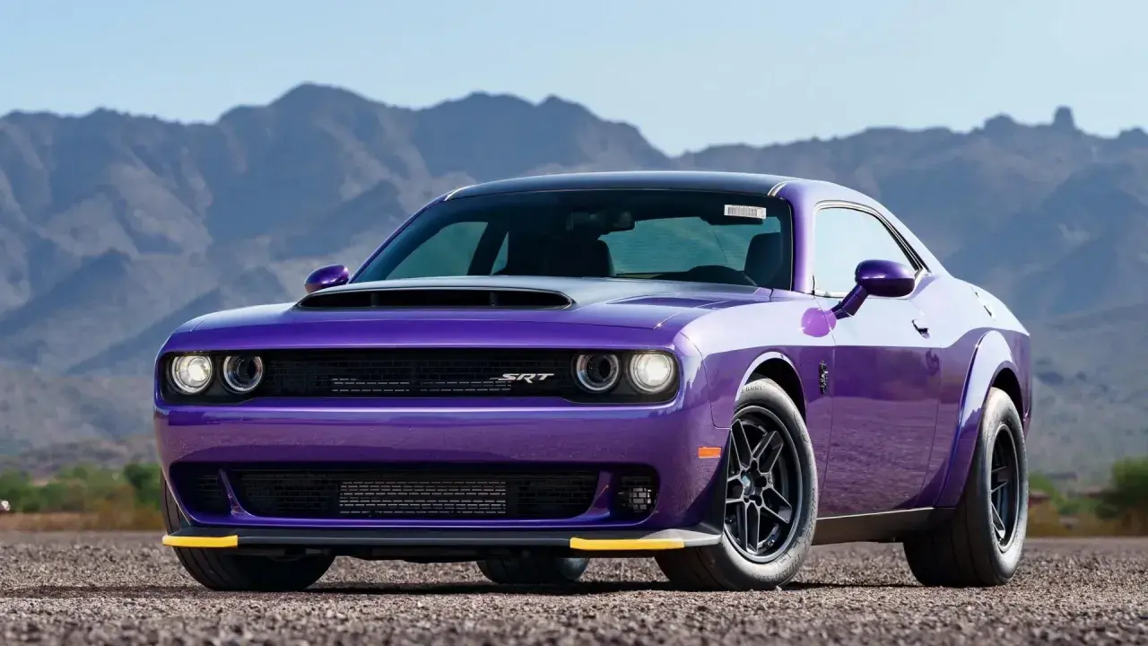 En lila amerikansk sportbil, en Dodge Challenger SRT, står parkerad på en grusväg med berg i bakgrunden.