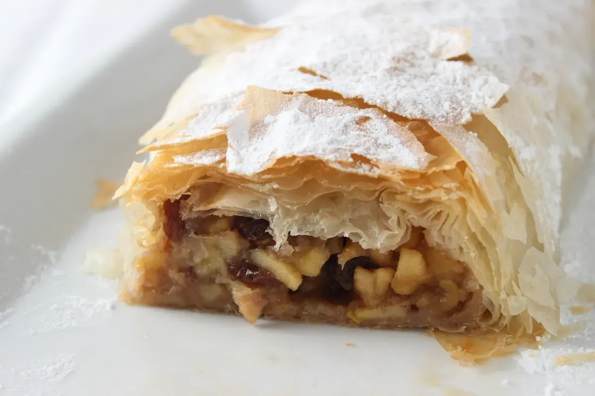 Un trozo de strudel de manzana, con su masa hojaldrada y relleno de manzana y pasas, espolvoreado con azúcar glas.
