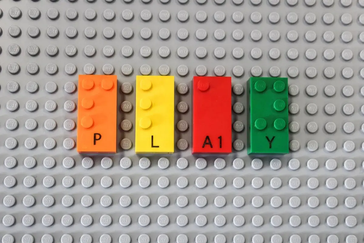Vier bunte LEGO-Steine bilden das Wort "PLAY". Die Steine sind orange, gelb, rot und grün. Das Erkennen von LEGO-Steinen ist der erste Schritt zum Bauen.