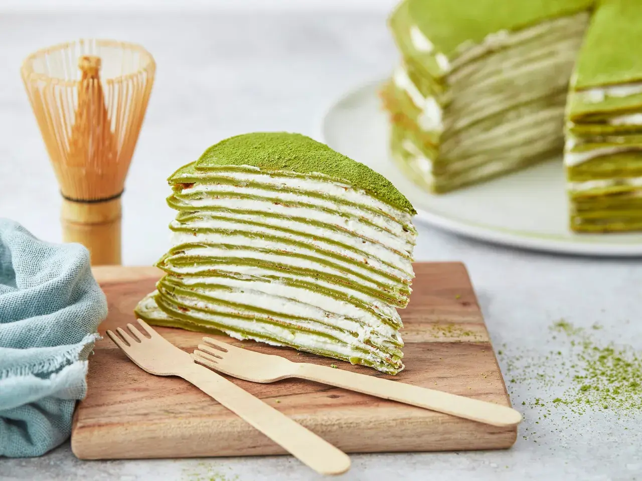 Ein Stück saftiger Matcha Kuchen mit vielen dünnen Schichten und cremiger Füllung, bestäubt mit grünem Pulver. Daneben ein Chasen und Holzgabeln.