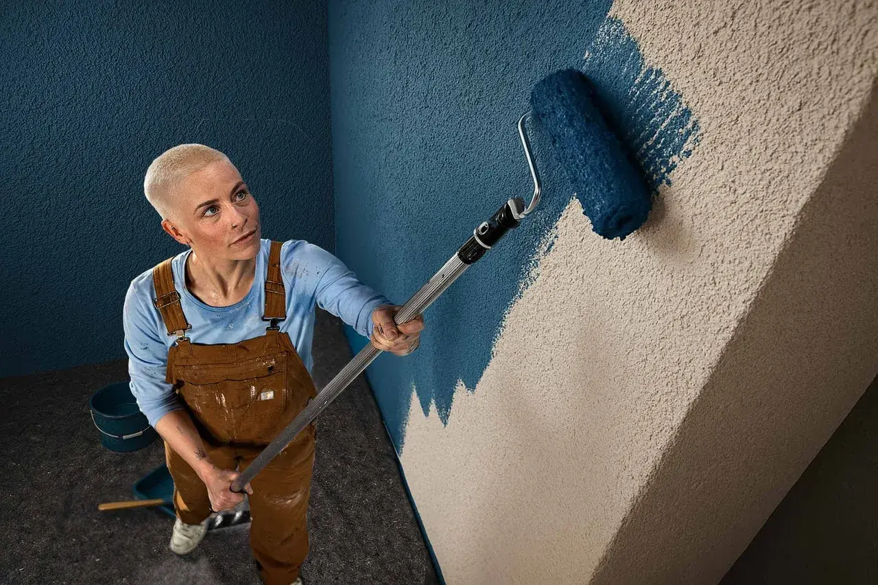 Frau mit Kurzhaarschnitt streicht mit einer Farbrolle eine Wand mit Rauputz. Sie trägt Latzhose und blaue Langarmshirt.