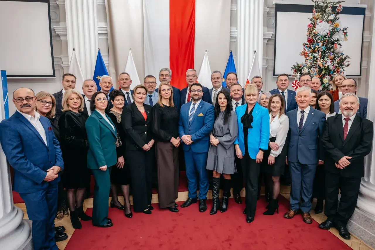 Grupa osób w eleganckich strojach pozuje do zdjęcia na tle flag i choinki.