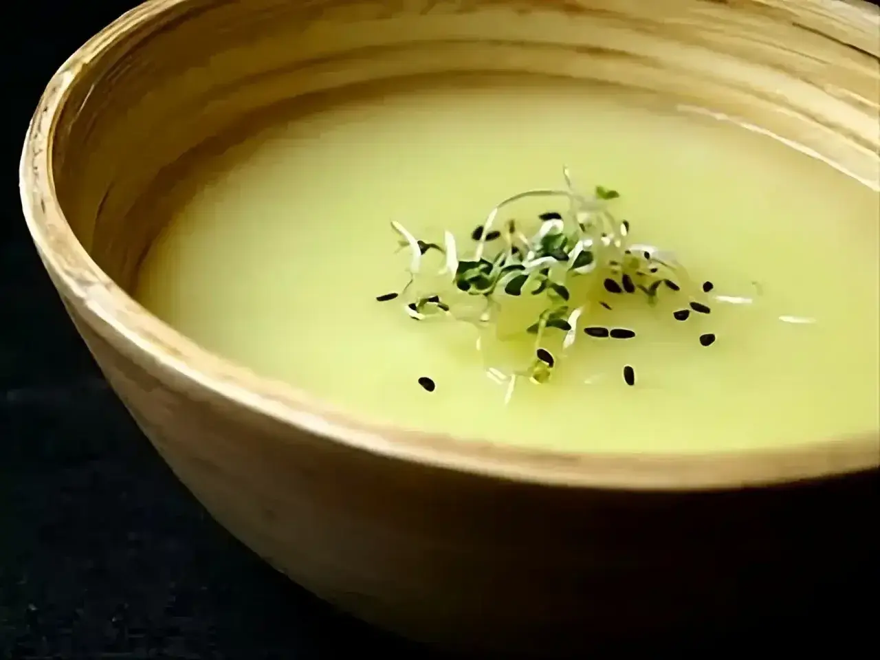 Un bol en bois contient un velouté d'asperges blanches, garni de pousses vertes et de graines noires.
