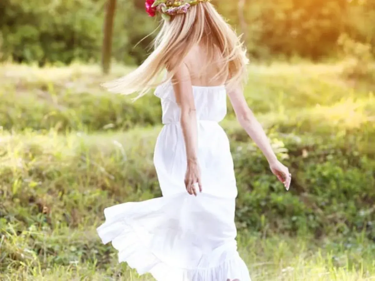 Une jeune femme aux cheveux blonds, portant une couronne de fleurs et une robe blanche légère, incarne l'esprit du printemps. Elle danse dans un champ verdoyant, une vision parfaite pour savoir comment s'habiller au printemps.