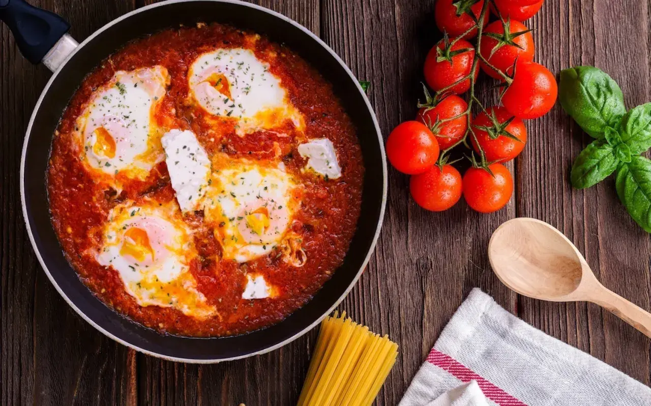 Uova al pomodoro in padella, con pomodorini freschi, basilico e un cucchiaio di legno, pronte per essere gustate.