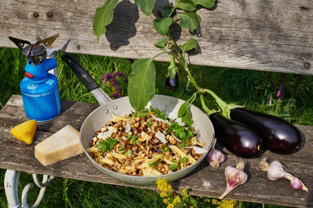 Pasta melanzane mit frischen Auberginen, Knoblauch und Parmesan. Ein Campingkocher und Zitrone deuten auf ein Outdoor-Mahl hin.