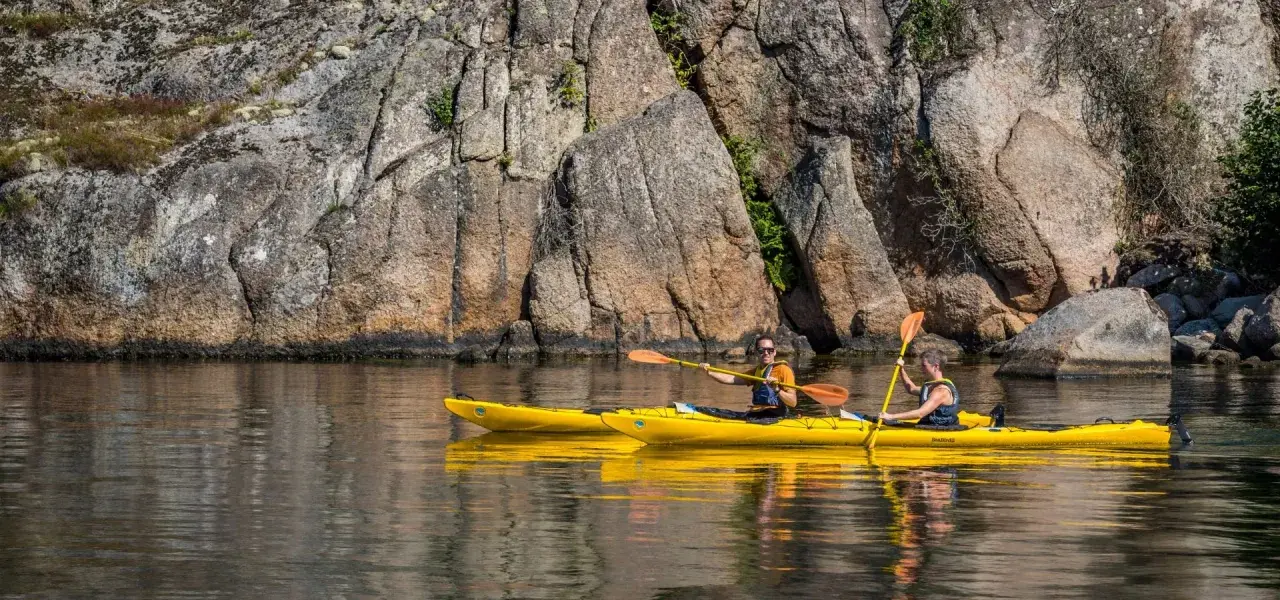 Två personer paddlar varsin gul kajak längs en klippig kust. En härlig dag för upplevelser i Sverige.