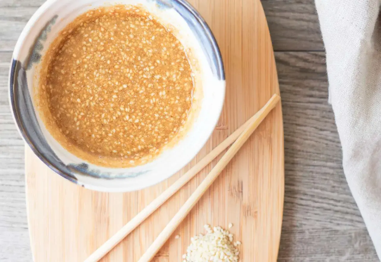 A bowl of creamy sesame sauce, perfect for your next sesame sauce recipe, sits on a wooden board with chopsticks and a sprinkle of sesame seeds.