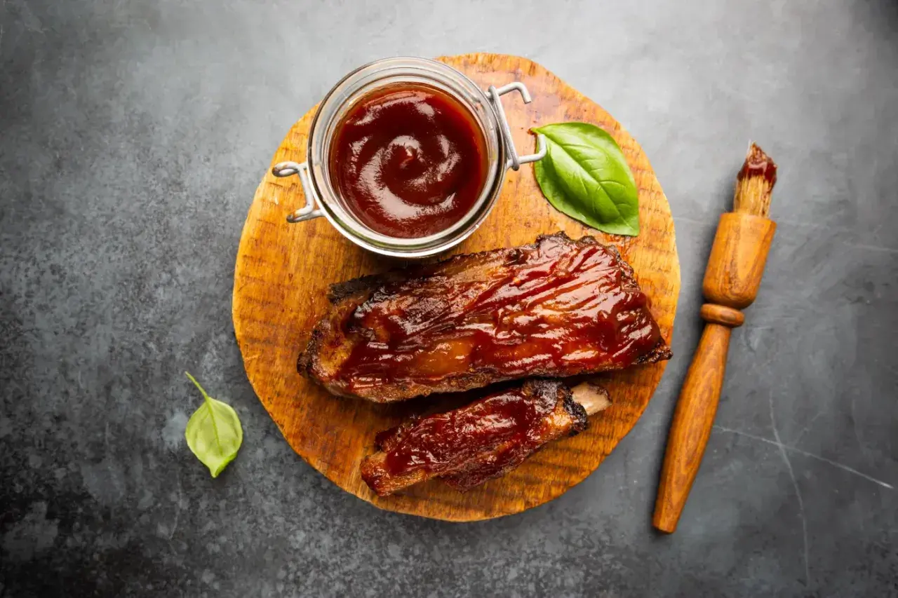 Soczyste żeberka w sosie barbecue, gotowe do podania. Obok słoiczek z sosem do steka i pędzelek.