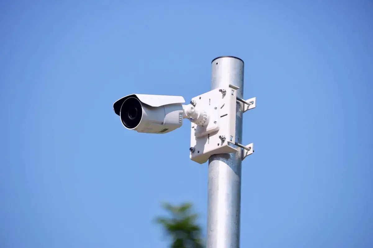 Biała kamera zewnętrzna zamontowana na metalowym słupku, idealna do monitorowania otoczenia. Dowiedz się, jak zamontować kamerę zewnętrzną.