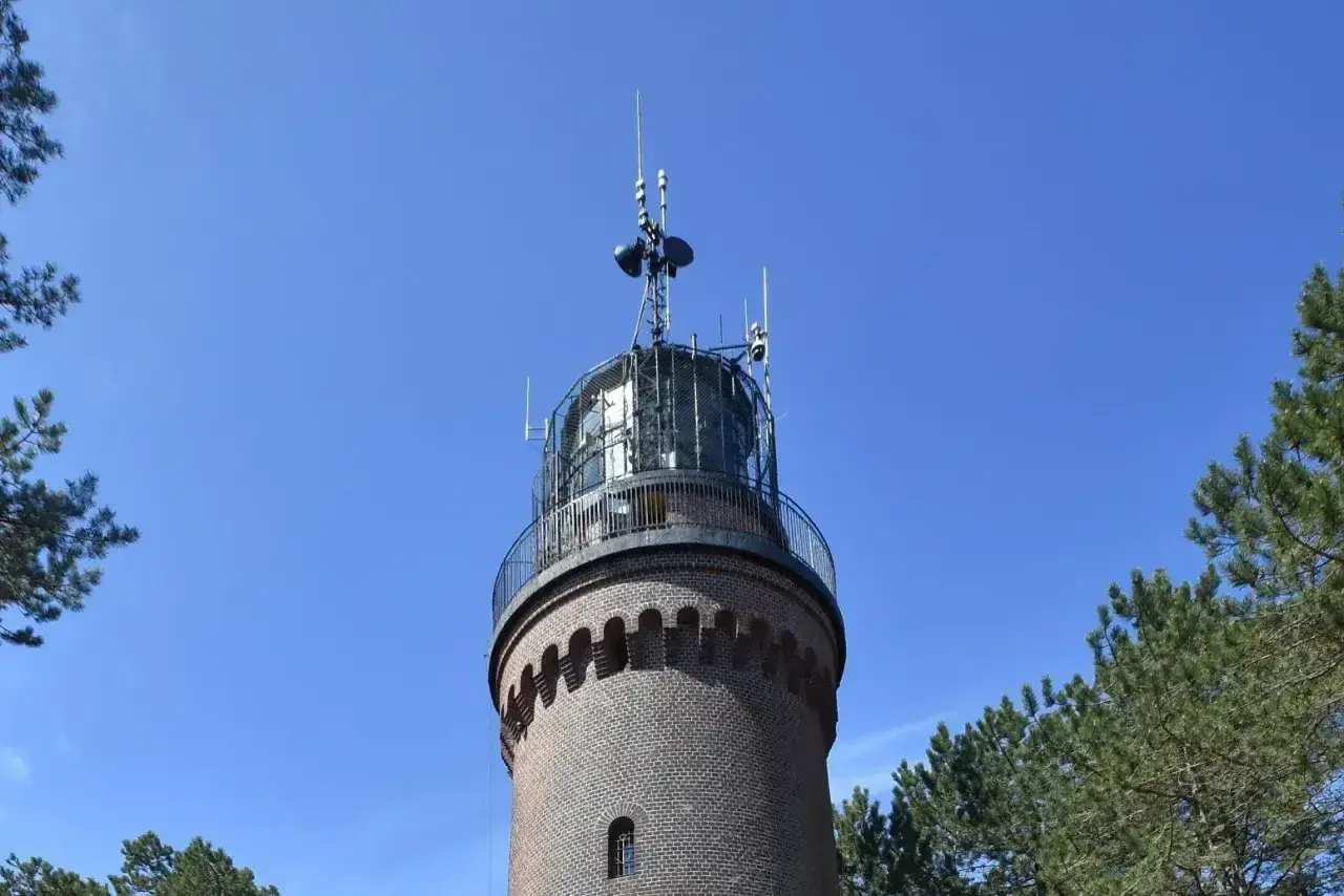 Ceglana łeba latarnia morska z metalową galeryjką i antenami na szczycie, otoczona sosnami, na tle błękitnego nieba.