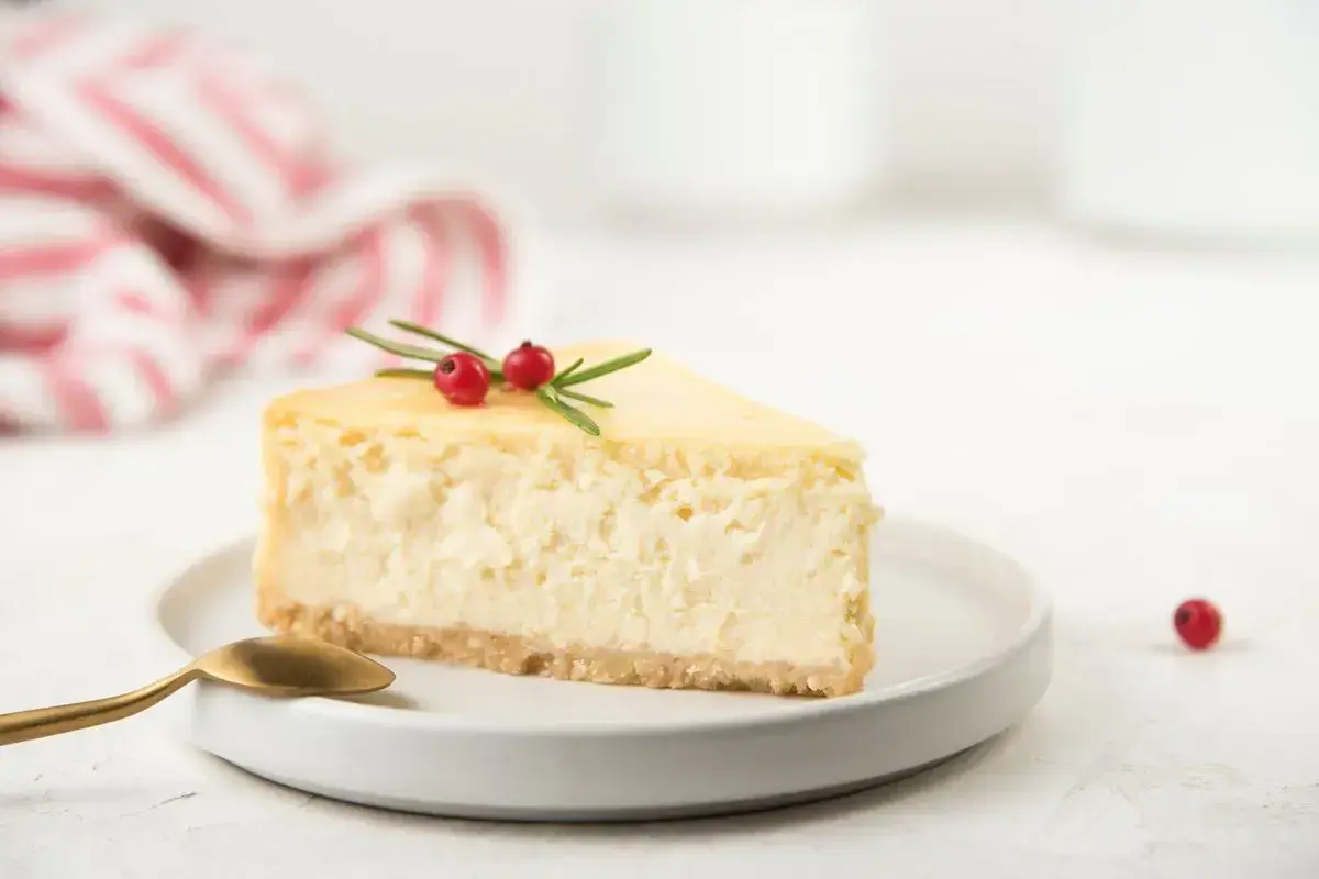 Una fetta di cheesecake con yogurt greco, guarnita con rosmarino e bacche rosse, su un piatto bianco con cucchiaino dorato.