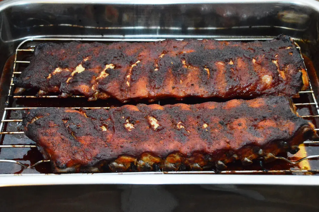 Zwei Rippchen-Stränge, dunkelbraun und saftig, ruhen auf einem Rost in einer Bratpfanne. Sie sind perfekt gegart, wie lange Spareribs im Ofen sein sollten.