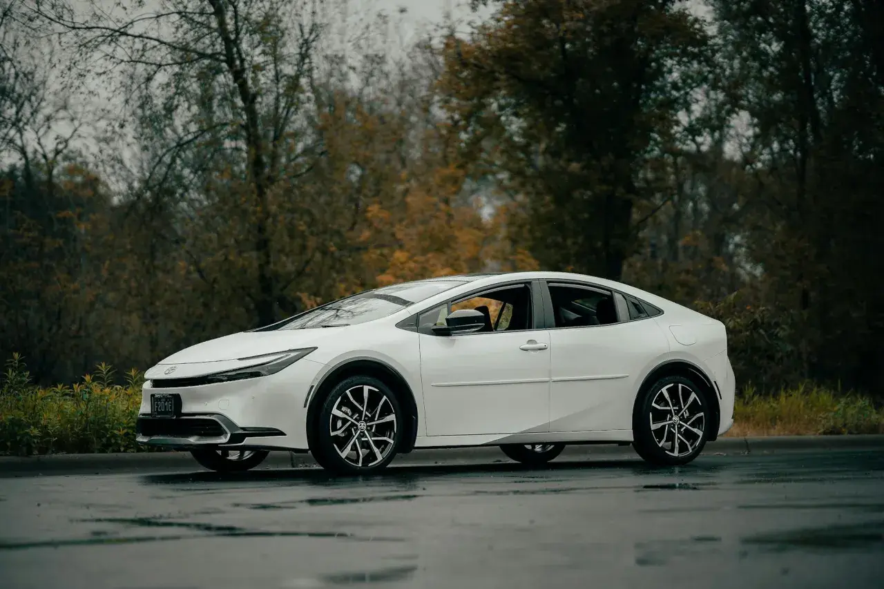 Biała Toyota Prius zaparkowana na mokrej drodze, z drzewami w tle.