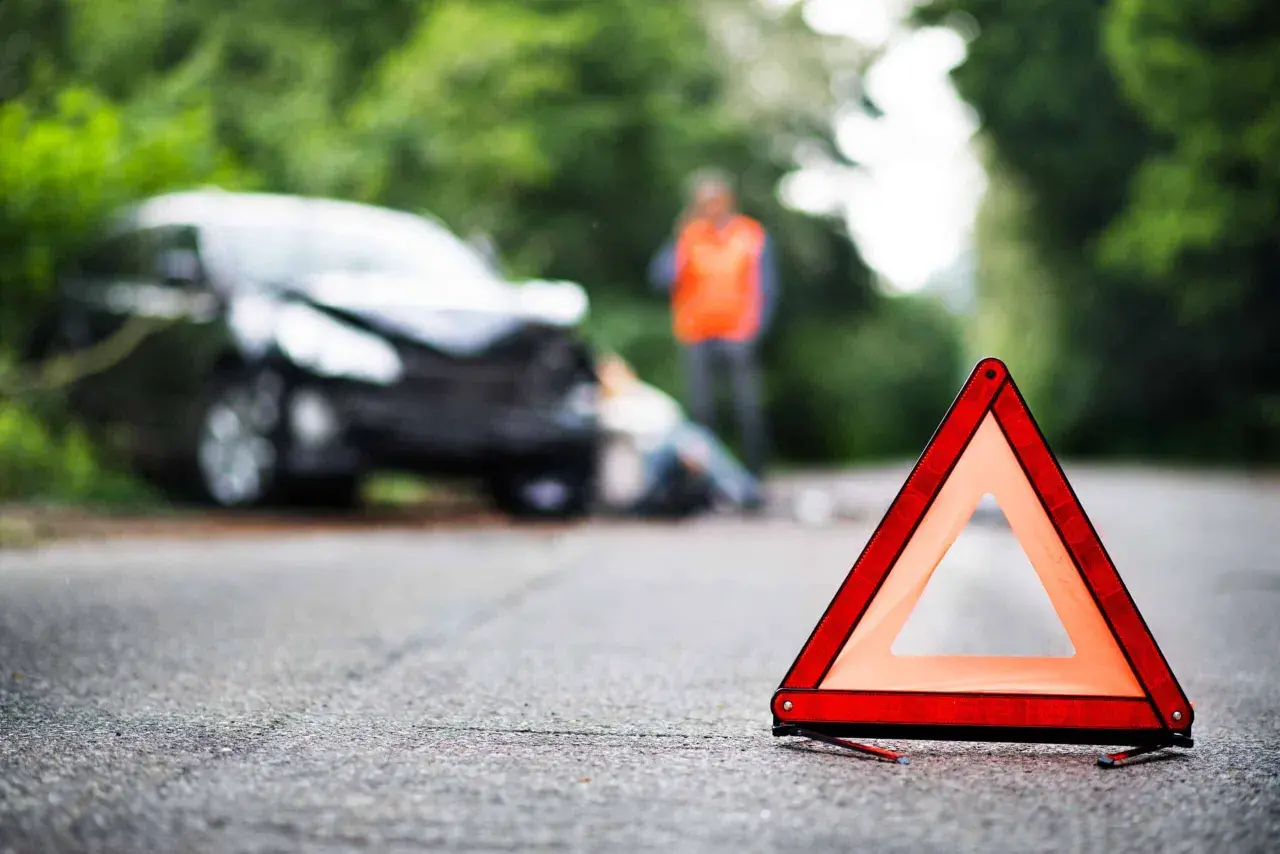 Triangle de signalisation sur la route, près d'une voiture accidentée où une personne gît au sol. Un homme en gilet orange se tient debout. Scène évoquant un homicide involontaire.
