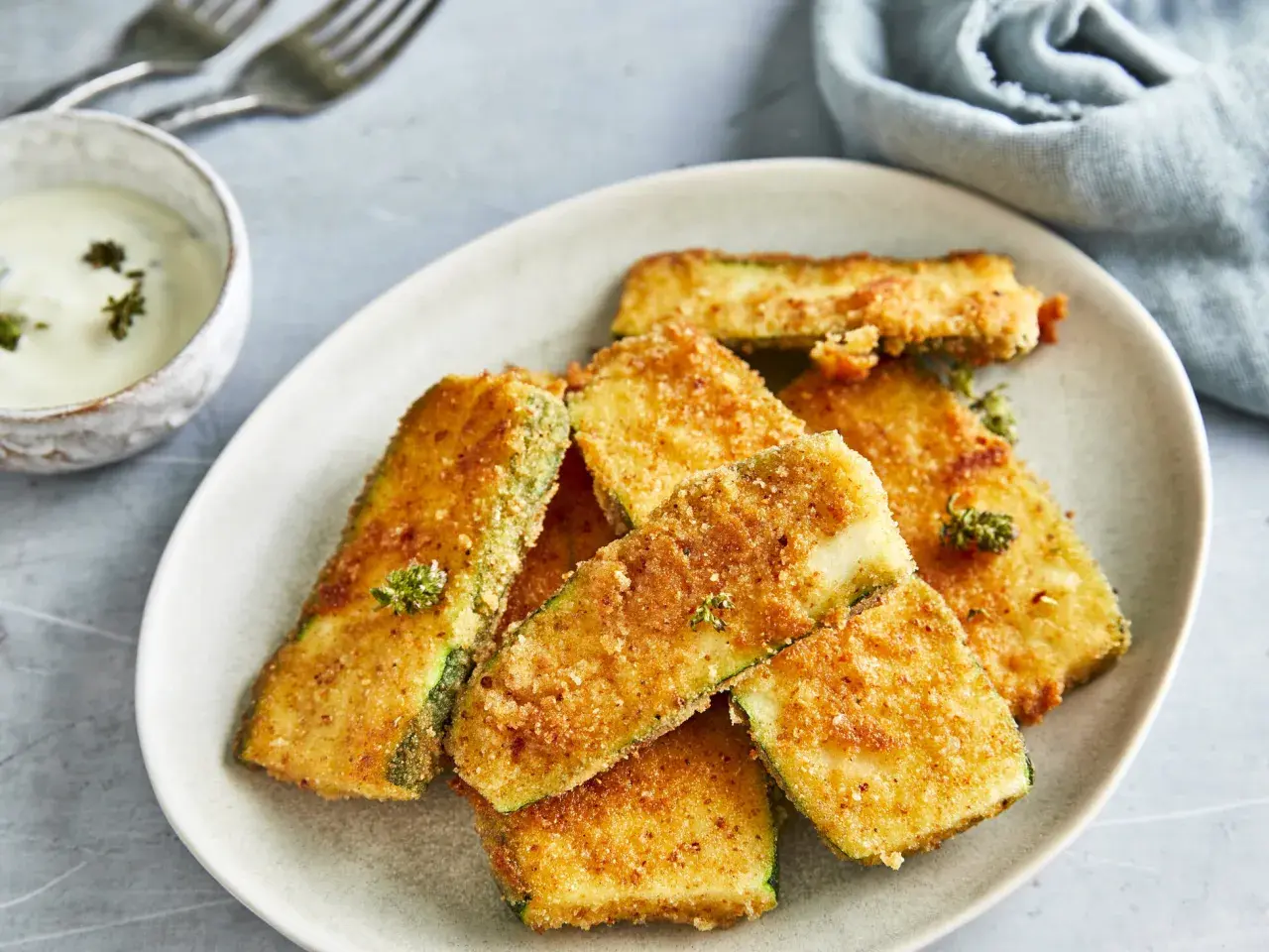 Knuspriges Zucchini-Schnitzel auf einem Teller, daneben eine Schale mit Dip und Gabeln.