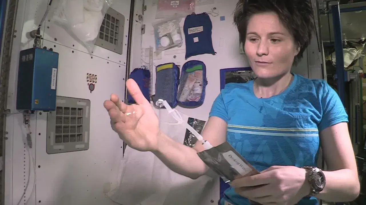 Astronautka pokazuje kroplę wody w dłoni, która unosi się w przestrzeni kosmicznej. Trzyma też opakowanie z napisem "Drinking Water".