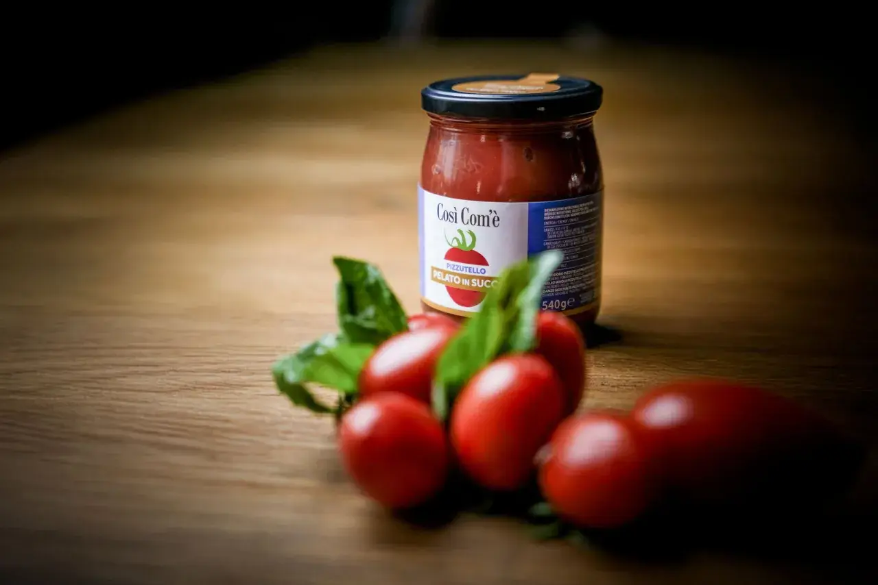 Barattolo di pomodoro siccagno pizzutello pelato in succo, con pomodorini freschi e basilico su un tavolo di legno.