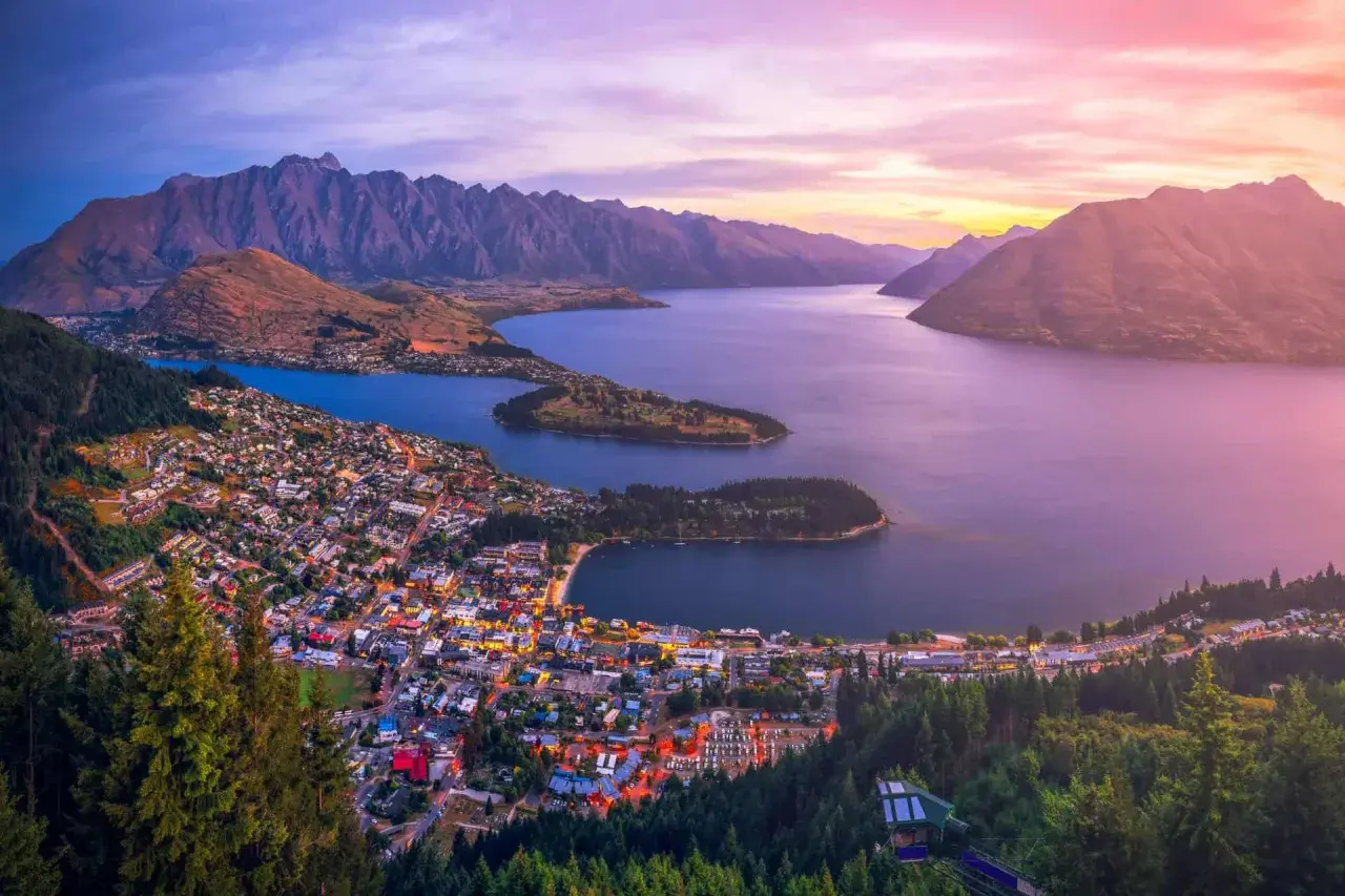 Vacker solnedgång över Queenstown, Nya Zeeland. Staden ligger vid en sjö, omgiven av berg och skog.