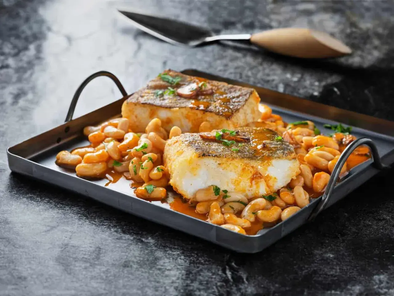 Dos lomos de bacalao a la llauna, dorados y jugosos, reposan sobre una cama de alubias blancas en salsa.